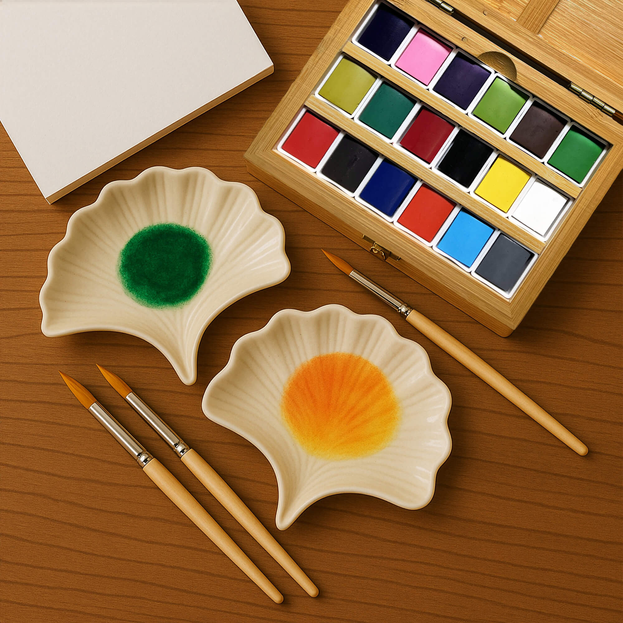 Watercolor paint set with two shell-shaped paint trays, brushes, and a wooden box on a wooden surface.