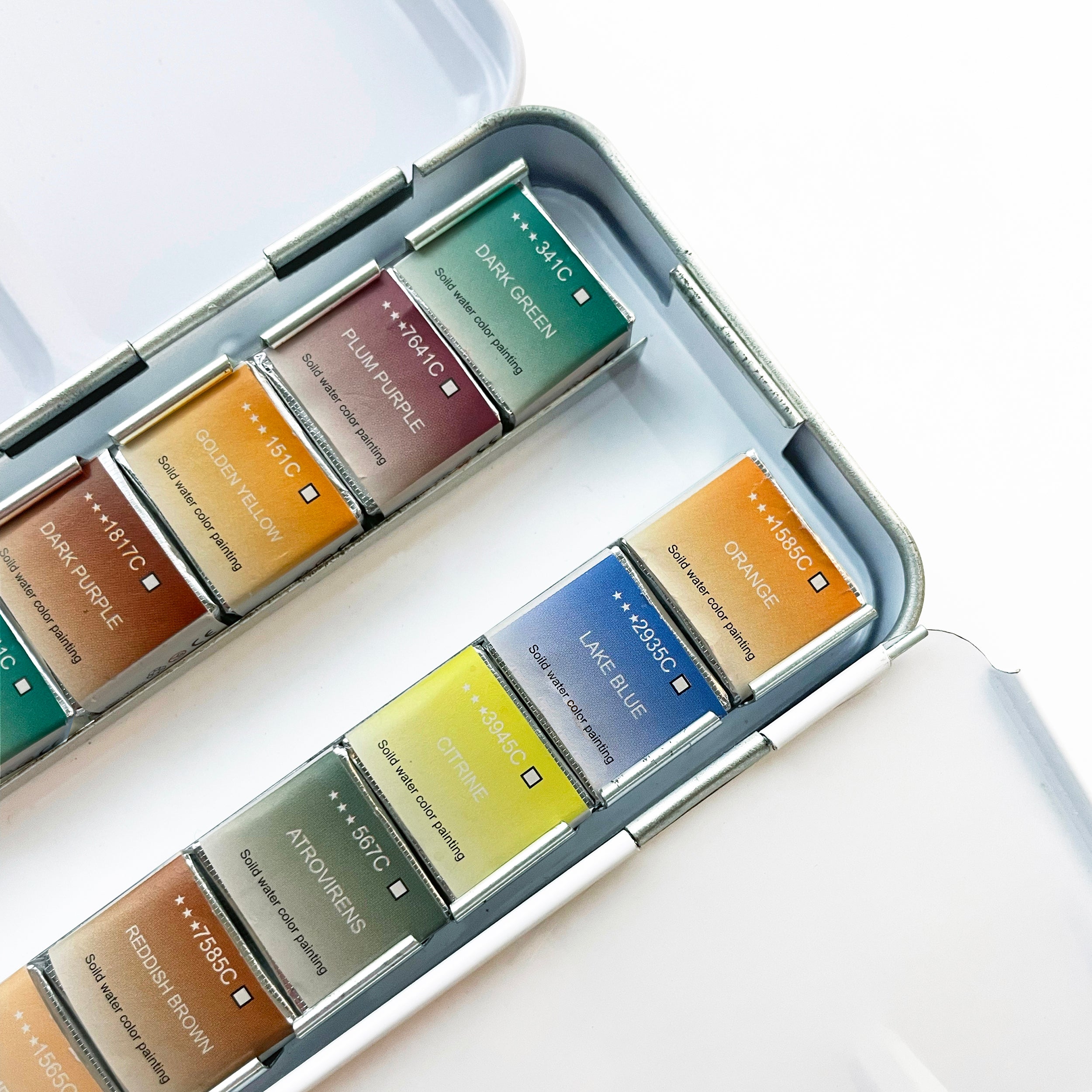 Close-up of the Elseware Unplug Watercolor Set in an aluminum travel case. The set features an array of vibrant solid watercolor paints, each wrapped in labeled paper indicating the color name and pigment code. The palette includes hues like dark green, plum purple, golden yellow, lake blue, citrine, and orange. Designed for artists of all levels, this eco-friendly set provides high-quality, sustainable colors for creative expression.