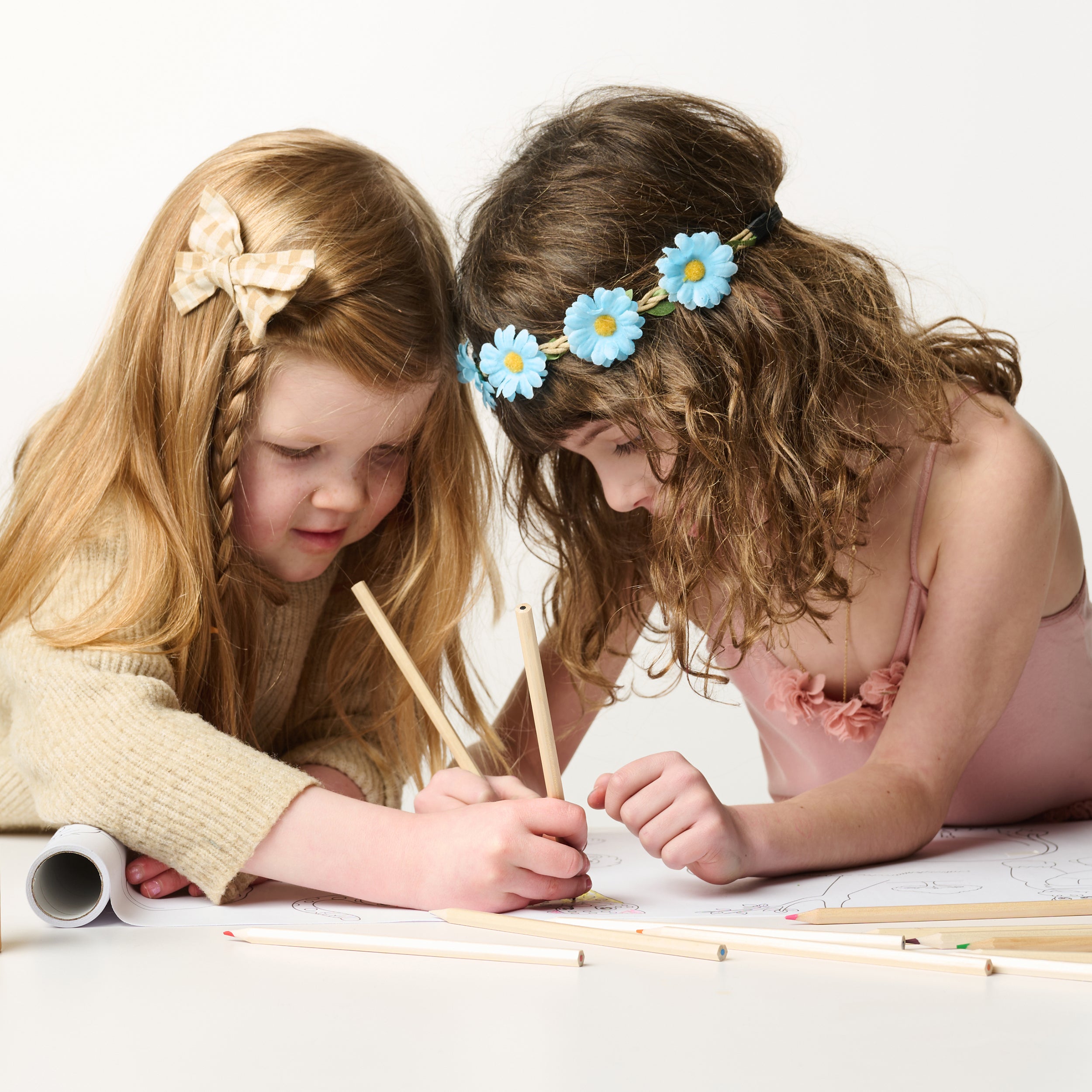 Two young girls coloring together with Elseware Unplug tall beechwood colored pencils on a large illustrated paper roll. One wears a beige sweater with a gingham bow, while the other wears a pink dress with a blue flower headband. Sustainable, non-toxic art supplies for creative play.