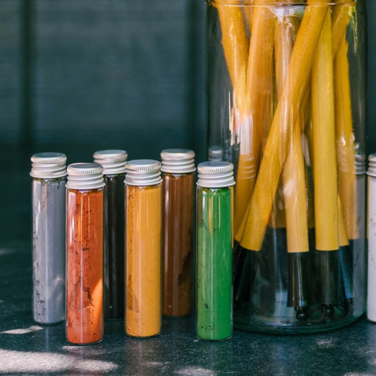 Elseware Unplug Earth Paint pigment vials arranged beside a jar of yellow paintbrushes, displayed in natural light on a dark surface.