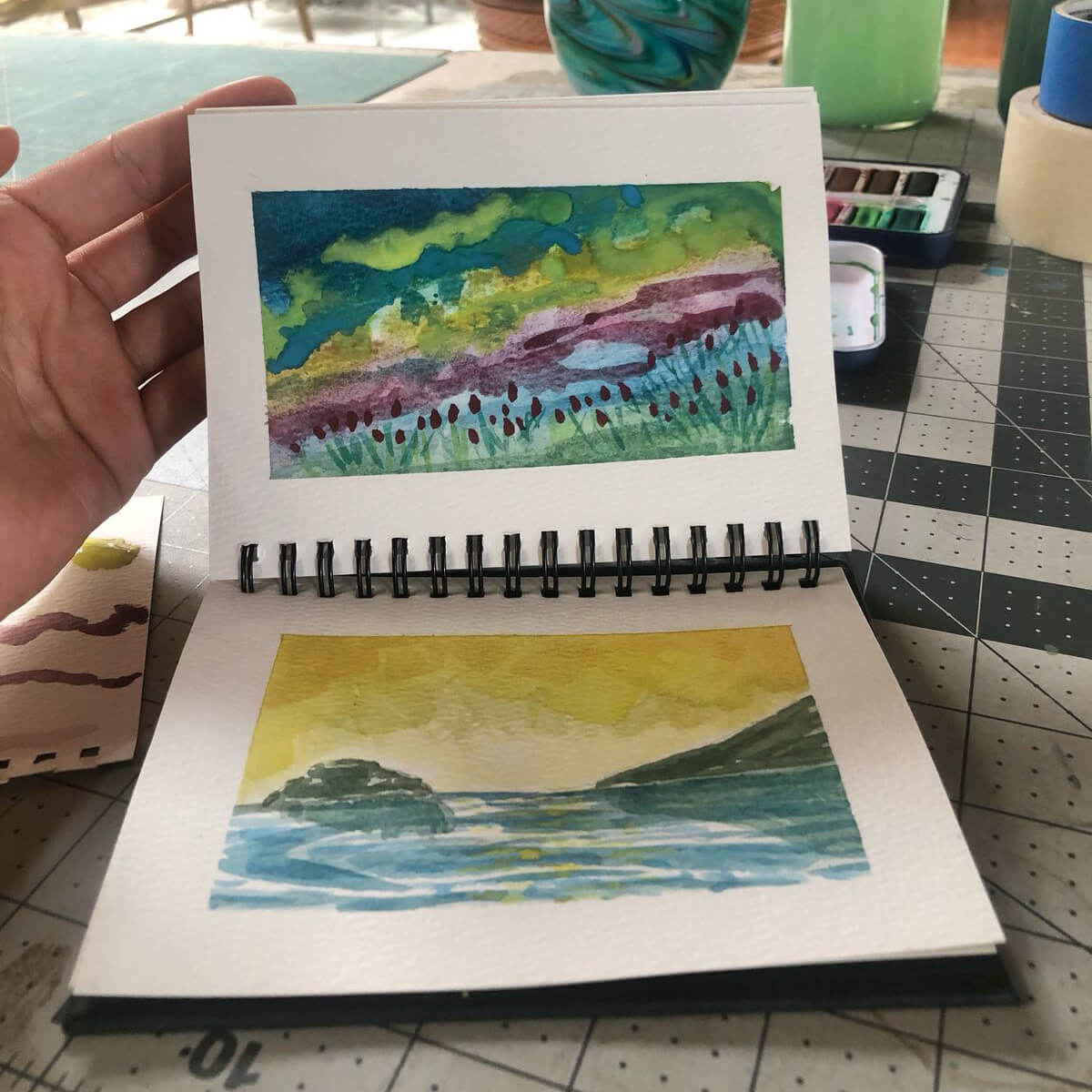 Open watercolor pad with colorful landscape paintings on textured paper, shown on a crafting table with art supplies nearby