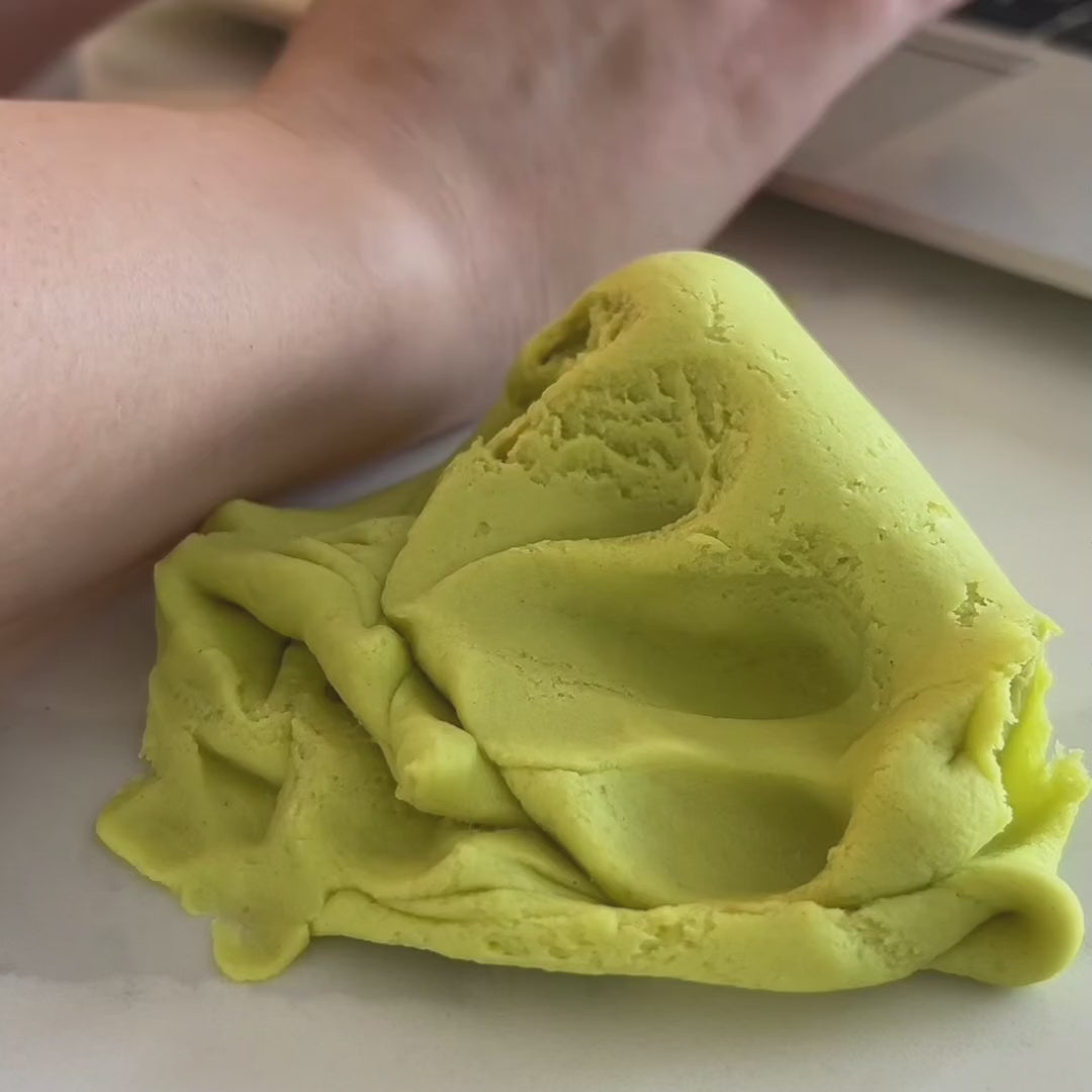 A close-up of Elseware Unplug's Mojito Therapy Dough, a soft, moldable dough in a refreshing green color, resting on a smooth surface. A person's arm is visible in the background, suggesting a moment of relaxation while using the dough. The texture appears airy and pliable, emphasizing its stress-relieving properties.