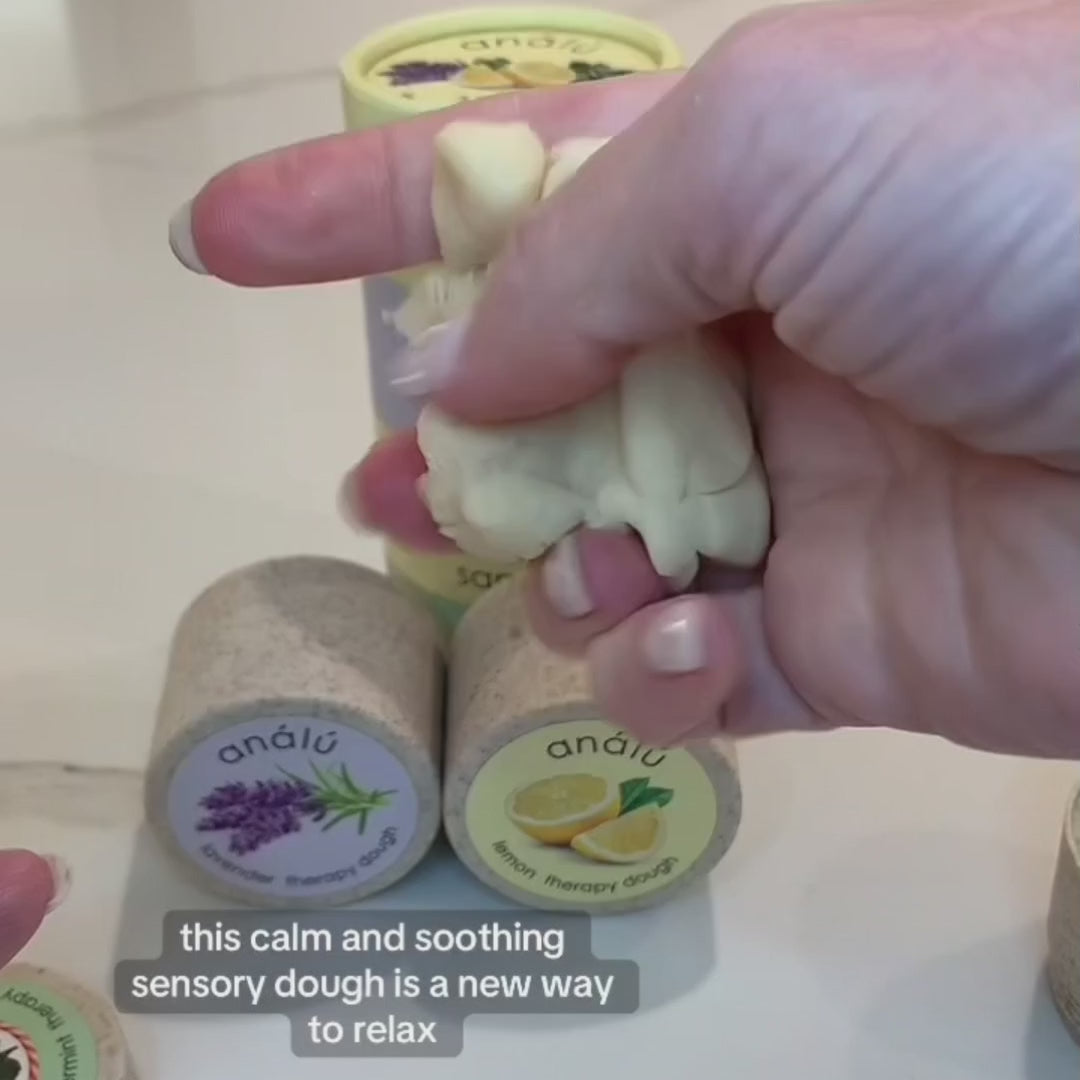 A hand squeezing soft, pale-colored therapy dough from the Análú Sampler set. In the background, two eco-friendly Análú therapy dough containers are visible, one labeled 'Lavender Therapy Dough' with a lavender illustration and the other labeled 'Lemon Therapy Dough' with a lemon illustration. A yellow cylindrical package with botanical artwork is partially visible. The caption on the video reads: 'This calm and soothing sensory dough is a new way to relax.