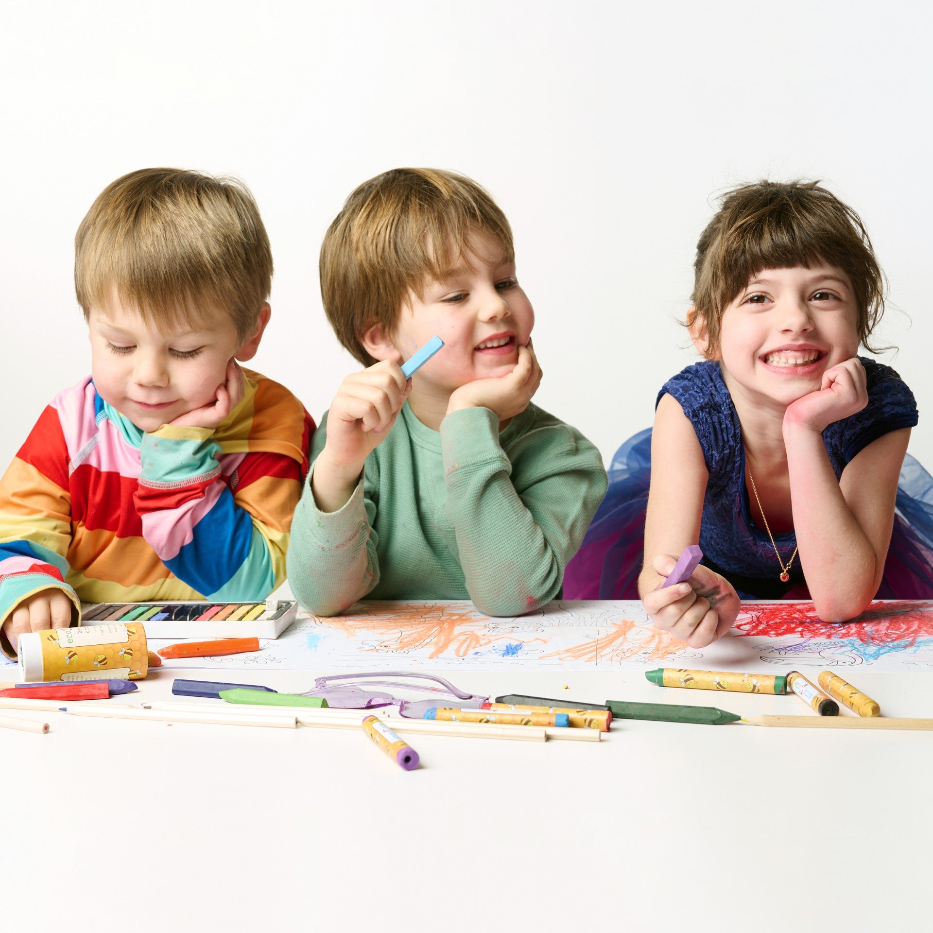 Three joyful children leaning on an Elseware Unplug Coloring Scroll, surrounded by non-toxic beeswax crayons, pastels, and colored pencils. The oversized paper roll is filled with colorful artwork, encouraging creativity, collaboration, and unplugged playtime.