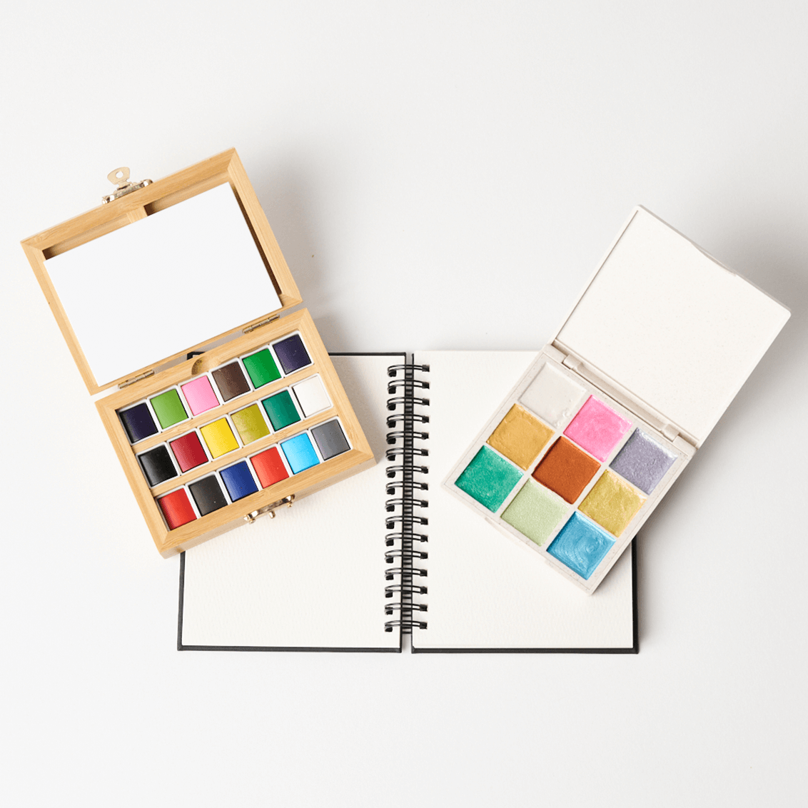 Two open watercolor palettes with a notebook on a white background
