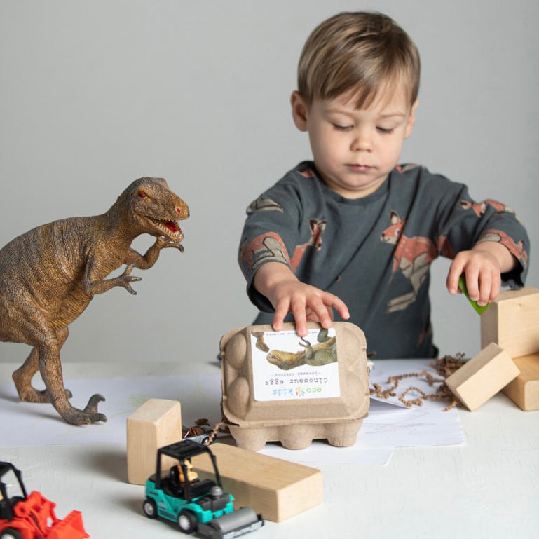 A young child playing with Elseware Unplug Dinosaur Egg Beeswax Crayons, housed in a recycled egg carton. The scene features wooden blocks, toy construction vehicles, and a realistic dinosaur figurine, creating a fun, imaginative play environment. These non-toxic, handmade crayons are shaped like dinosaur eggs, making coloring a prehistoric adventure.