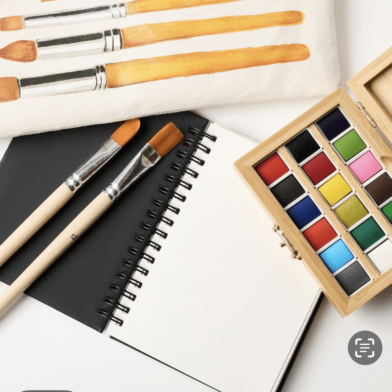 Set of paintbrushes, watercolor paints, and a notebook on a white surface