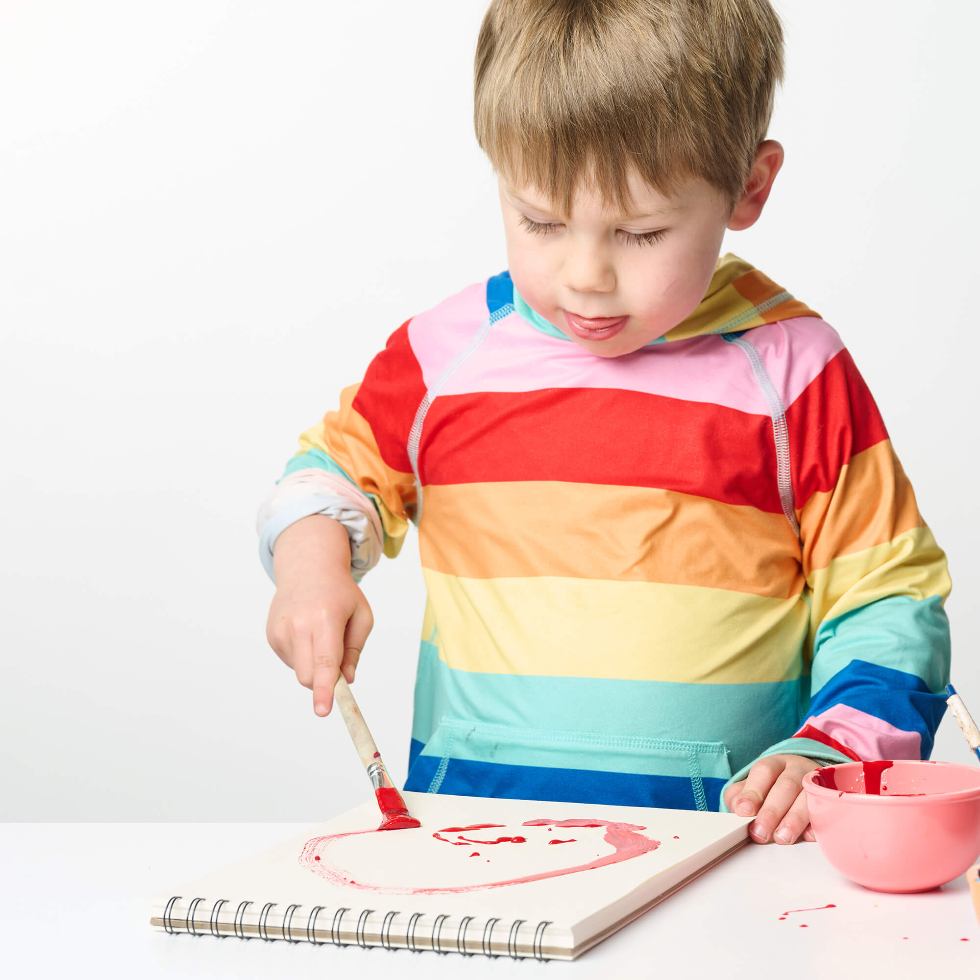Young child in a rainbow-striped shirt painting with a red brush on a spiral-bound Elseware Unplug sketch pad.
