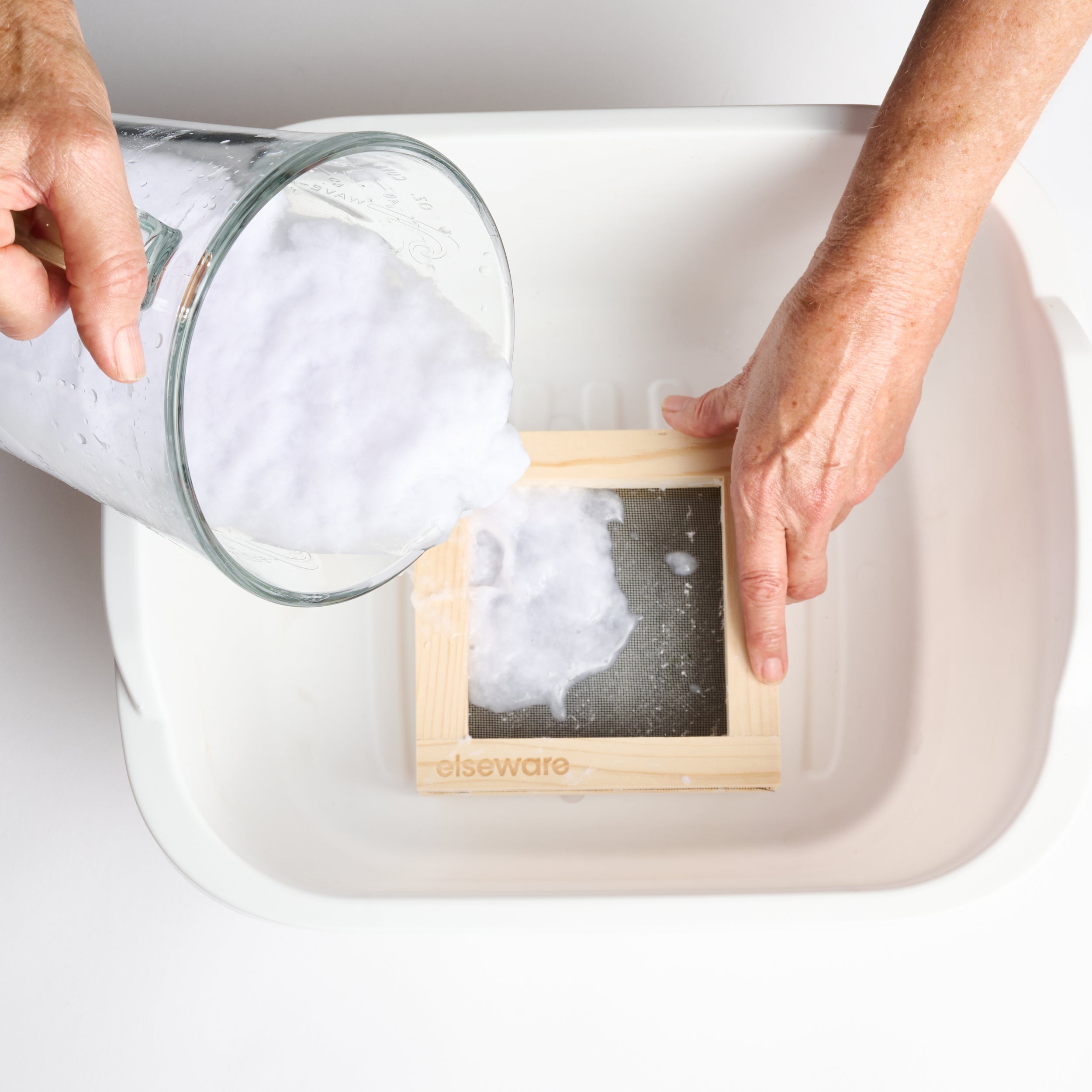 Hands pouring recycled paper pulp onto an Elseware Unplug wooden paper-making frame, set inside a water basin. This eco-friendly Paper Making Kit includes a pine wood mold and deckle, sponge, and herbal inclusions for added texture and color. A simple, rewarding craft for children and adults to create handmade paper with love.