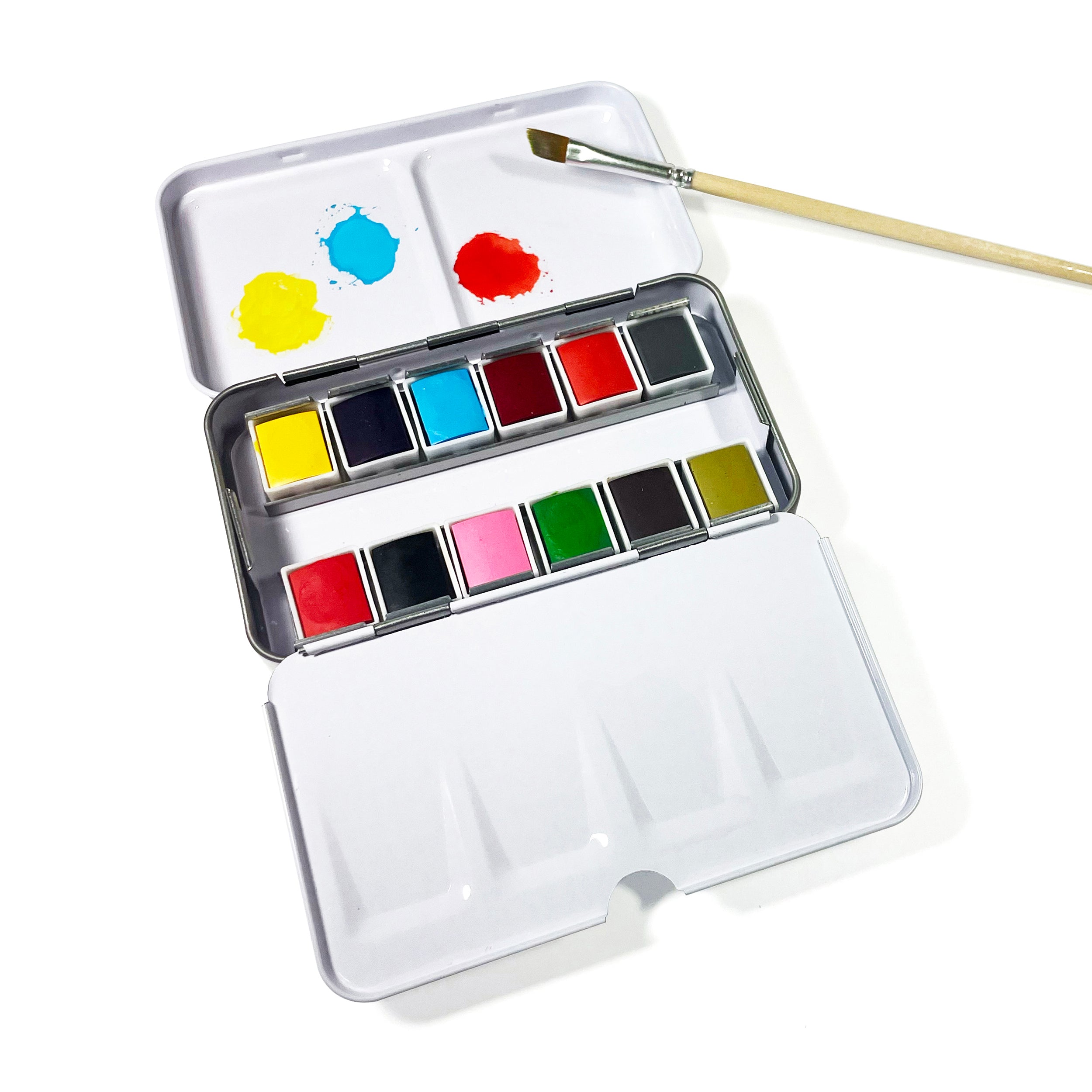 Elseware Unplug Watercolor Set in an open aluminum travel case, displaying 12 vibrant watercolor pans in a variety of colors. The lid serves as a built-in mixing palette with splashes of blue, yellow, and red paint. A wooden-handled paintbrush rests above the case, ready for use. This eco-friendly set is designed for easy portability and creative exploration.