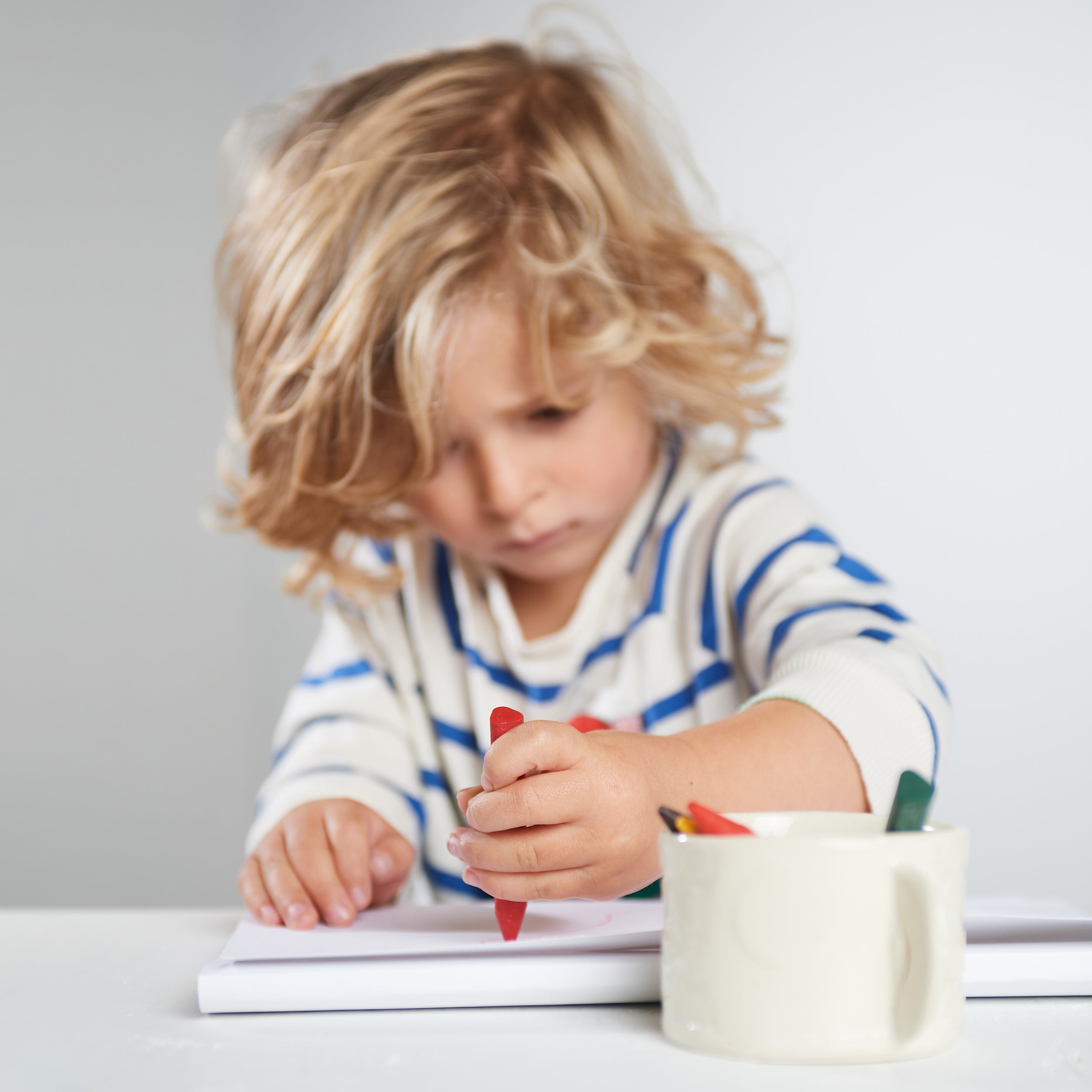 Young child with blonde curly hair wearing a striped blue and white shirt, drawing with a red Elseware Unplug triangle-shaped beeswax crayon on white paper. A ceramic cup holds additional non-toxic, natural beeswax crayons in yellow and green.