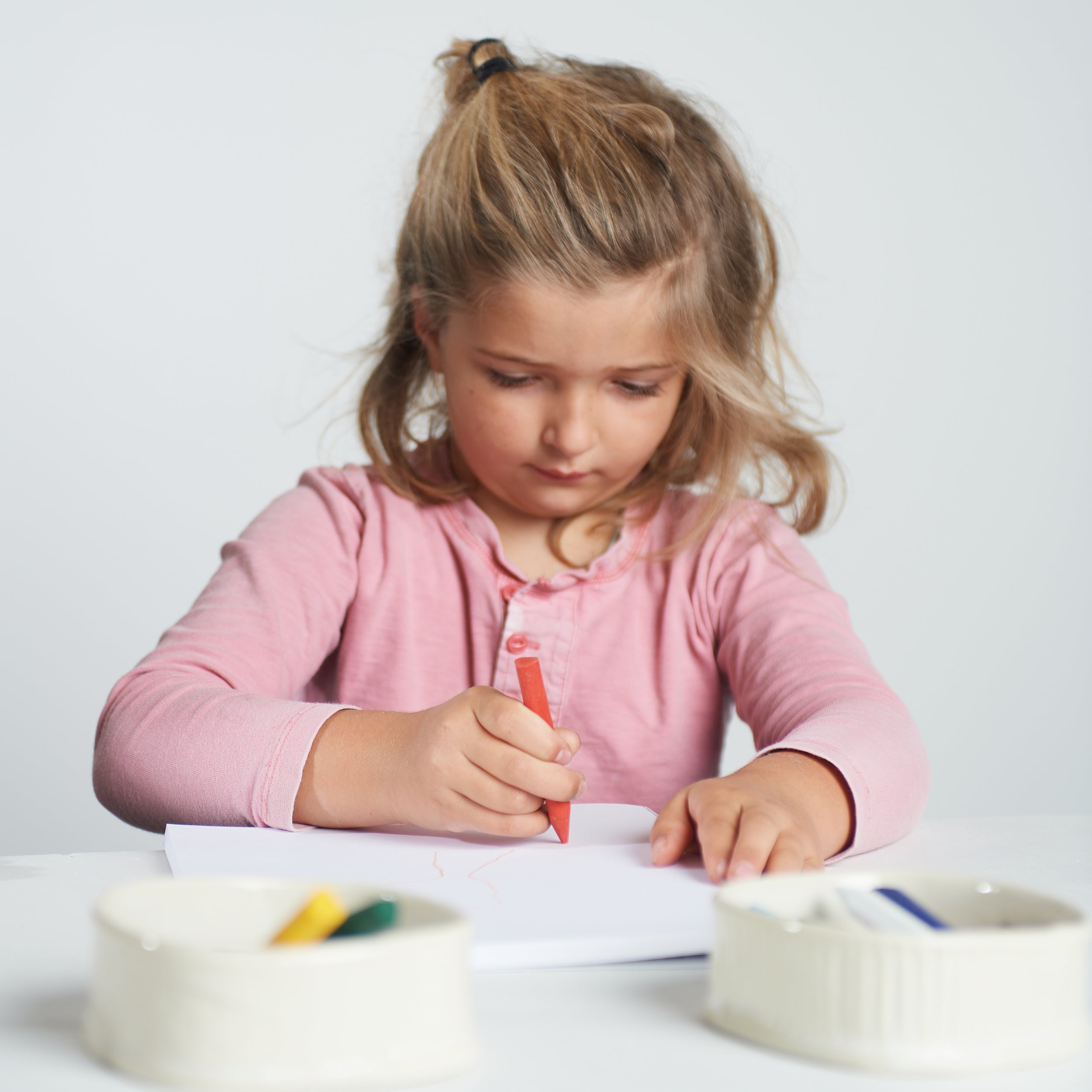 Young child in a pink long-sleeve shirt drawing with a red Elseware Unplug triangle-shaped beeswax crayon on white paper. Non-toxic, natural beeswax crayons in yellow and green are visible in a ceramic dish nearby.