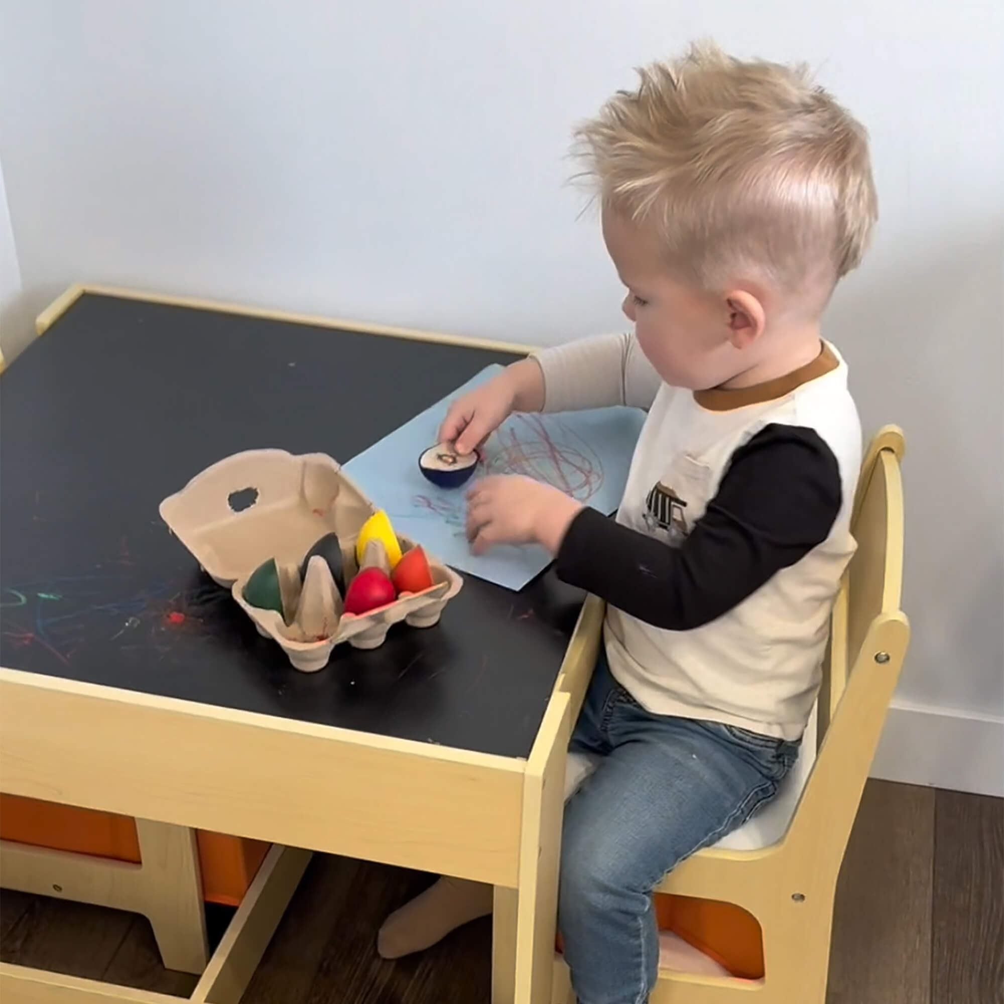 Young child coloring with Elseware Unplug’s Dino Egg-Shaped Beeswax Crayons at a small wooden table. The open egg carton displays vibrant, easy-to-hold crayons designed for little hands.