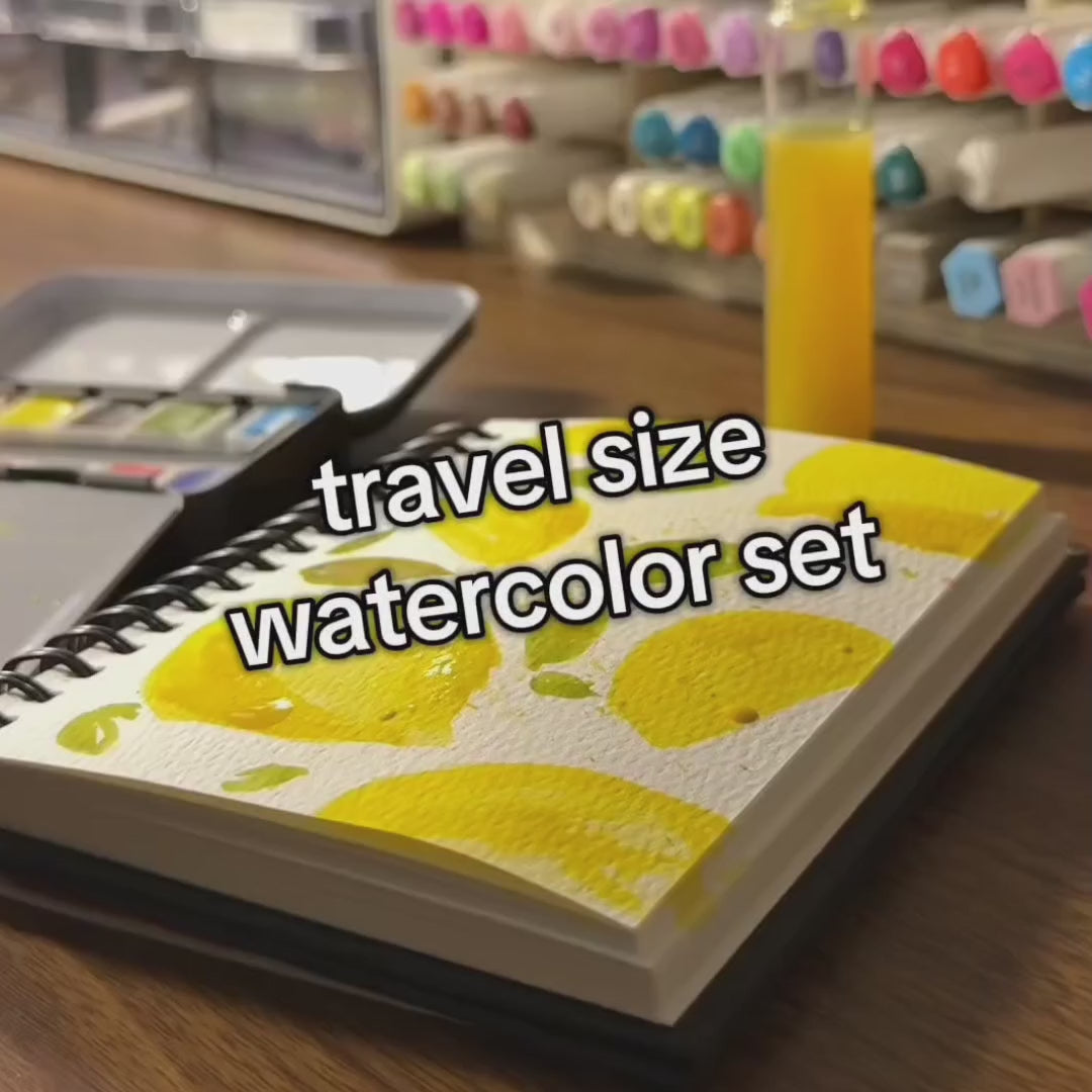 Video of a portable Watercolors To Go Kit in action. An artist paints vibrant strokes on the included paper pad, showing off the rich pigment and smooth texture. The compact travel case is opened to reveal neatly organized watercolor pans, a brush, and sponge—perfect for creating on the go.
