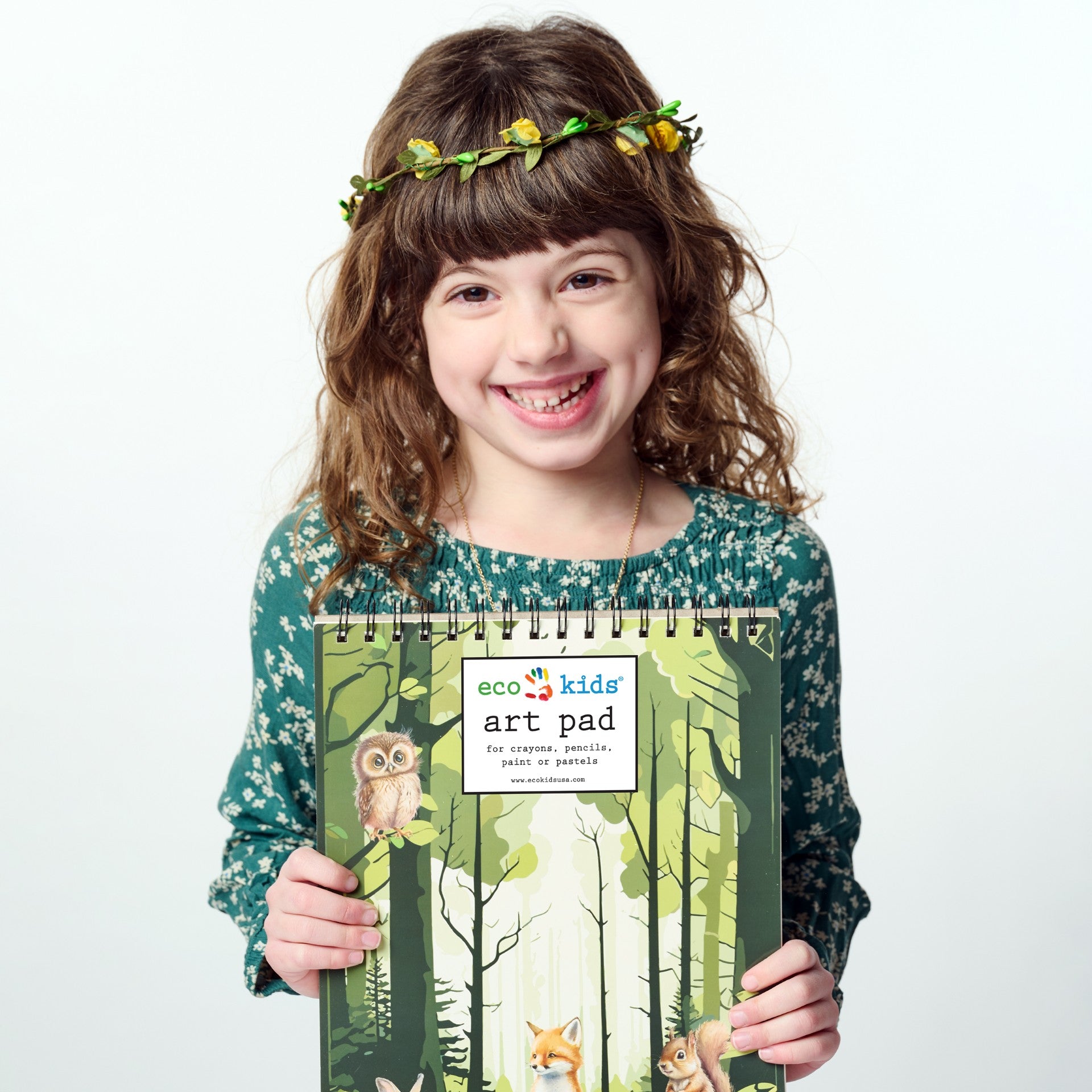 Smiling young girl wearing a green floral dress and a yellow flower crown holding the Elseware Unplug 8.5” x 11” Forest Art Pad. This spiral-bound drawing pad features 50 pages of smooth, high-quality paper, perfect for crayons, colored pencils, pastels, markers, and paint.