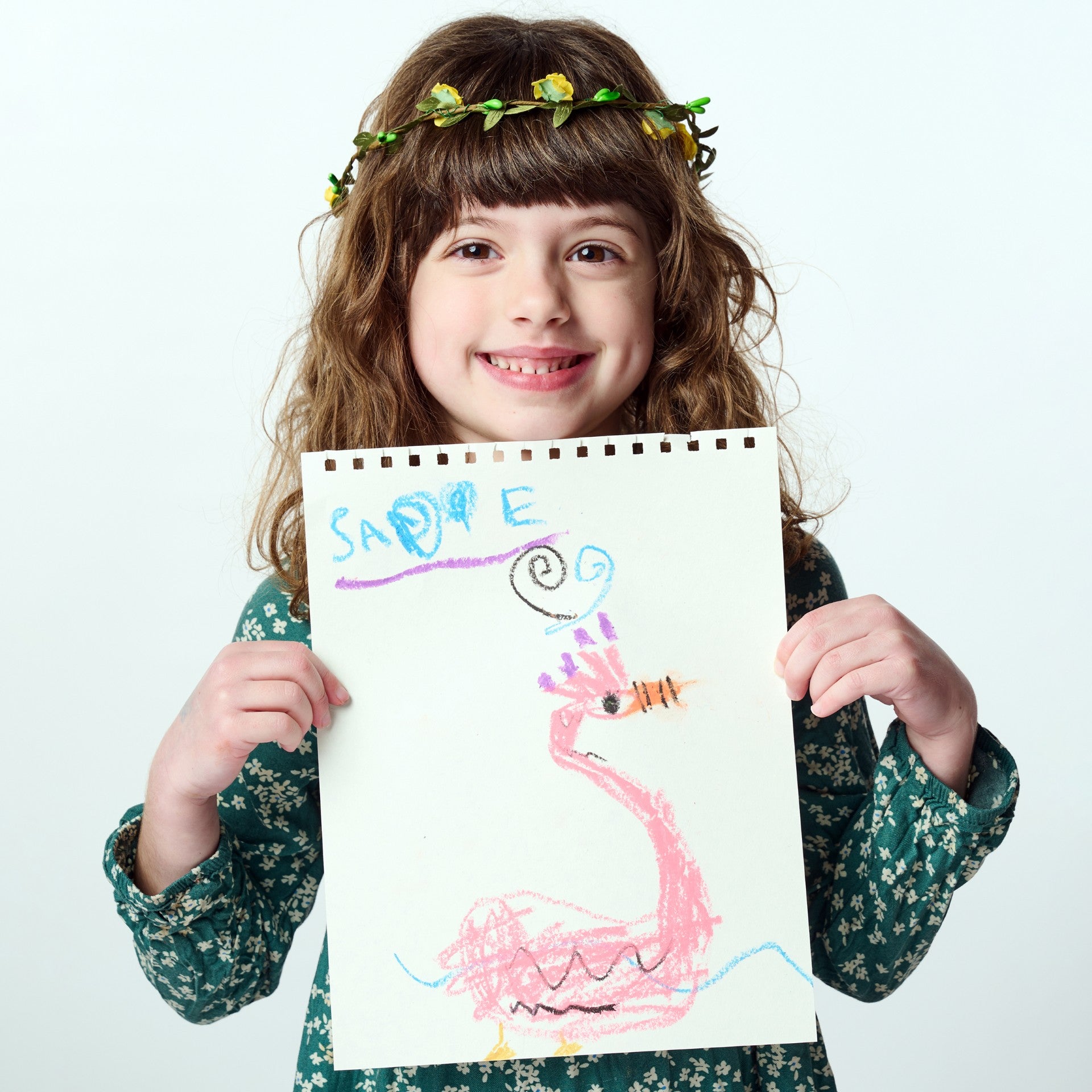 Young girl wearing a green floral dress and a yellow flower crown proudly holding up her artwork created on an Elseware Unplug 8.5” x 11” drawing pad. Featuring 50 pages of smooth, high-quality paper, this spiral-bound art pad is perfect for colored pencils, crayons, pastels, markers, and charcoal.