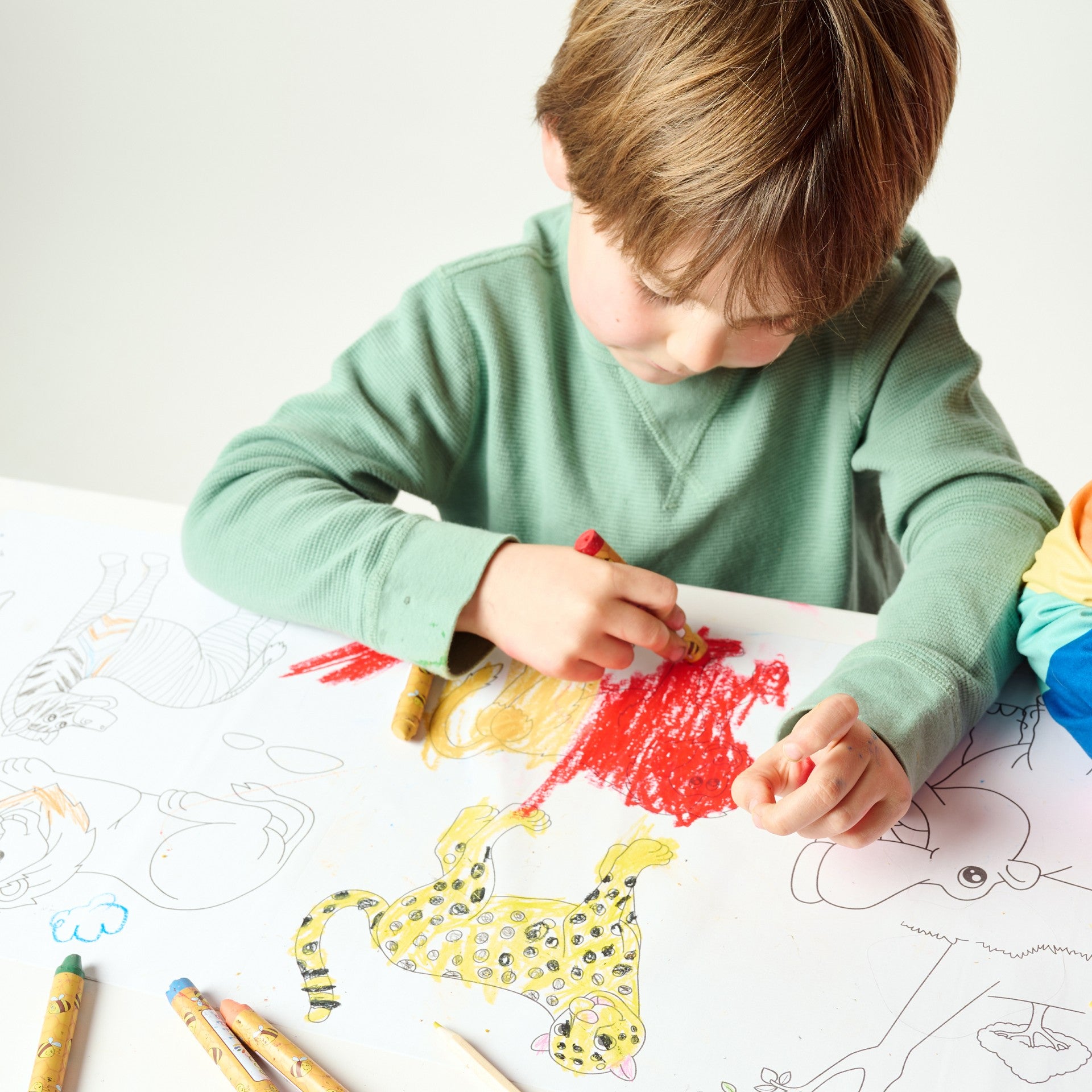 Young child in a green long-sleeve shirt coloring a jungle-themed Elseware Unplug Coloring Scroll with vibrant, non-toxic beeswax crayons. The oversized paper roll features playful animal illustrations, encouraging creativity and hands-on artistic fun.